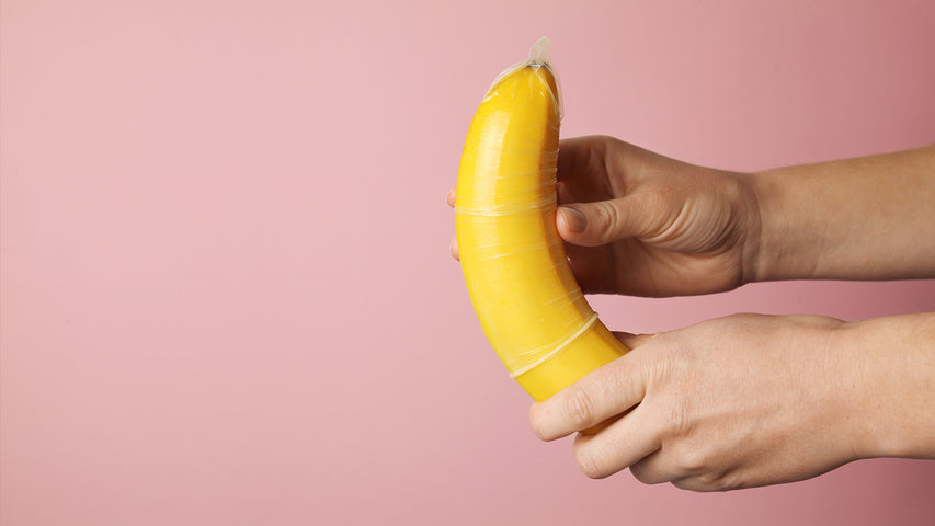 Hands putting a condom on a banana in front of a pink background.