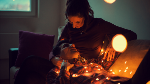 Woman and man laying on the sofa together while looking at each other and covered in holiday string lights.