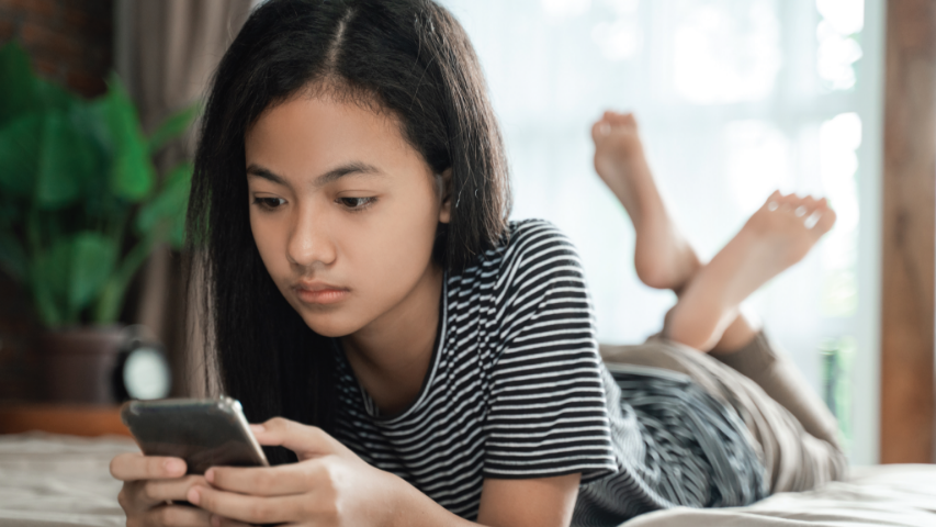 Young girl wearing a striped shirt laying on her stomach in bed while browsing on her cell phone.