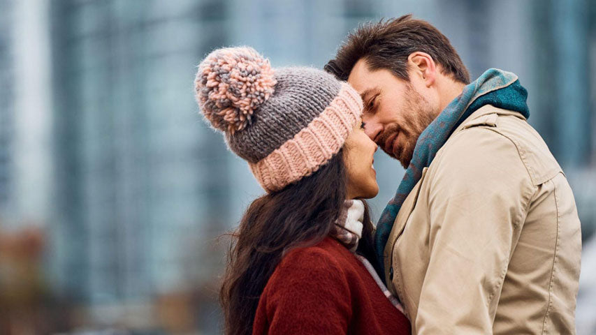 Woman in toque with a pompom going in to kiss her partner in an outdoor setting.