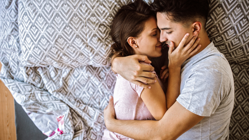 Two people holding each other tightly in bed on top of grey patterned pillowcases and bed sheets.