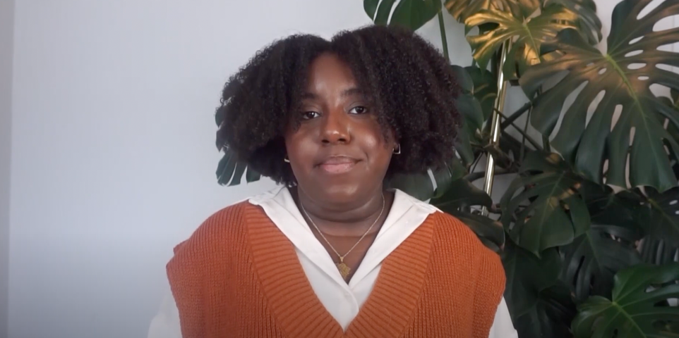 Girl wearing orange sweater vest and white shirt looking into the camera with a plant behind her