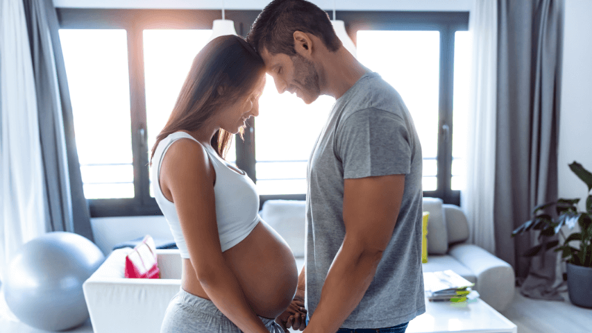 Une femme enceinte et son partenaire regardent le ventre arrondi de la femme dans un salon éclairé.