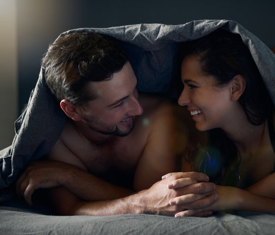 Couple interlocking fingers while smiling at each other under the bed sheets