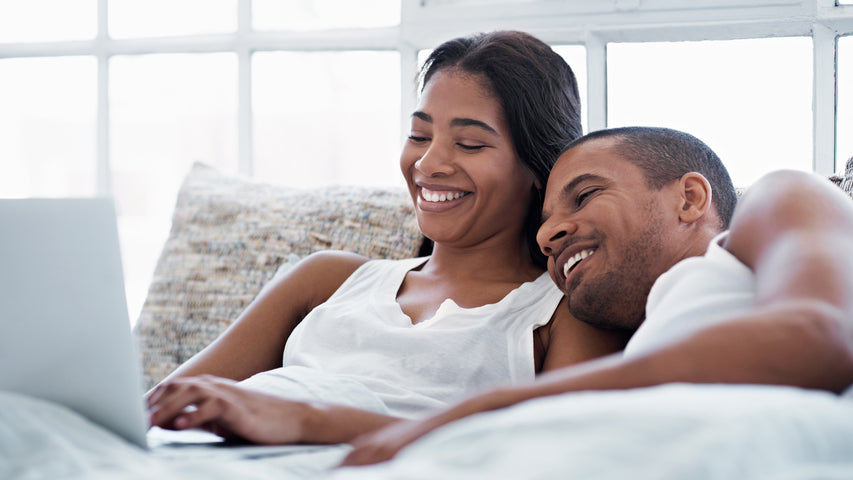 A smiling couple lies in bed watching a laptop.