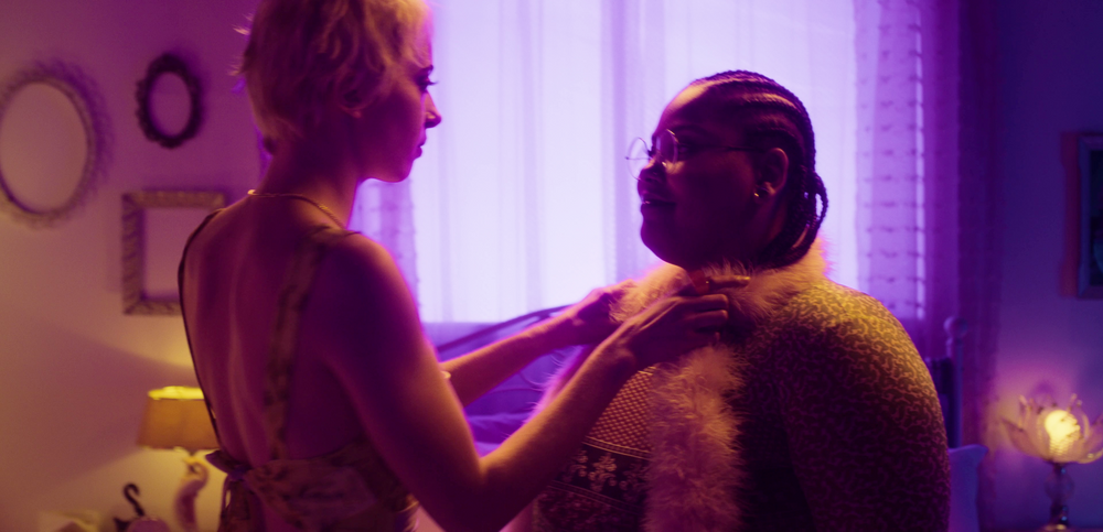Une femme qui tend les bras et met les mains sur les épaules de sa partenaire dans une chambre à l’éclairage violet.