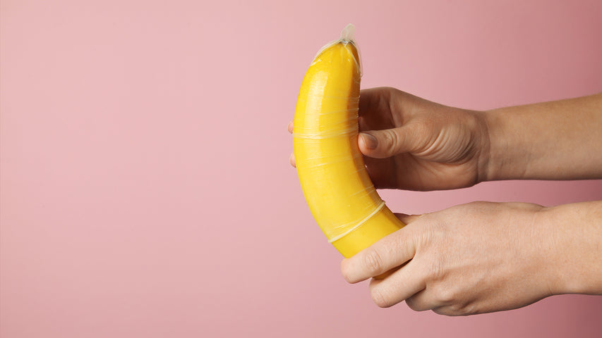 Hands putting a condom on a banana in front of a
pink background.