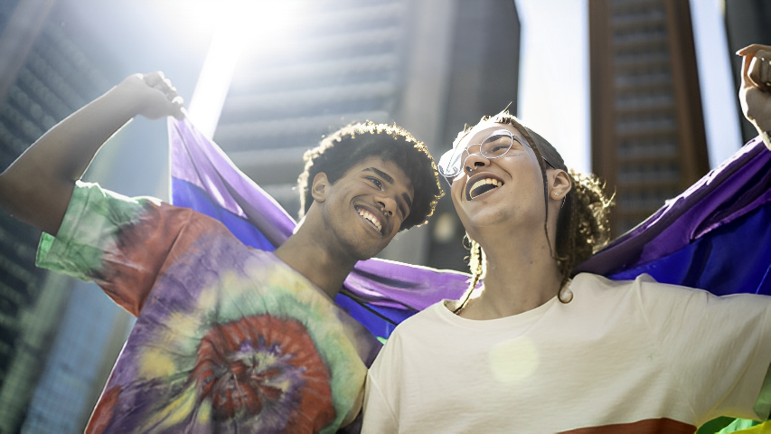 Two people smiling while holding a Pride flag behind them in front of skyscrapers.