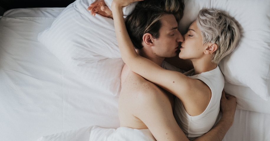 Couple kissing on top of white sheets in bed.