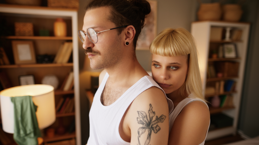 Two people wearing white tank tops staring off together in a room with two bookshelves and a lit lamp.