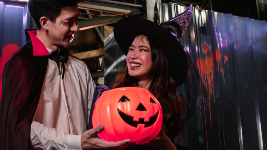 Man dressed as a vampire and woman dressed as a witch holding a jack-o’-lantern decoration smile at each other.