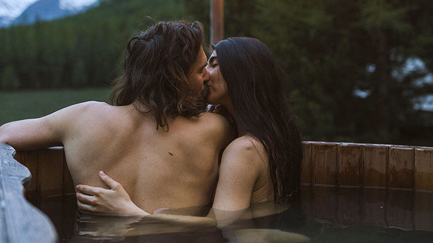 Two lovers sharing a kiss in a remote hot tub together next to hanging lights, a forest, and mountains.