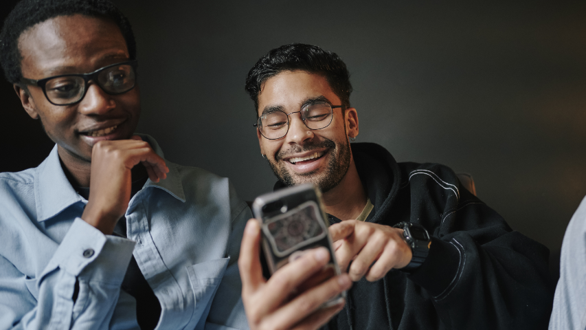 Two men sitting together, one showing something on his phone while the other watches with interest.