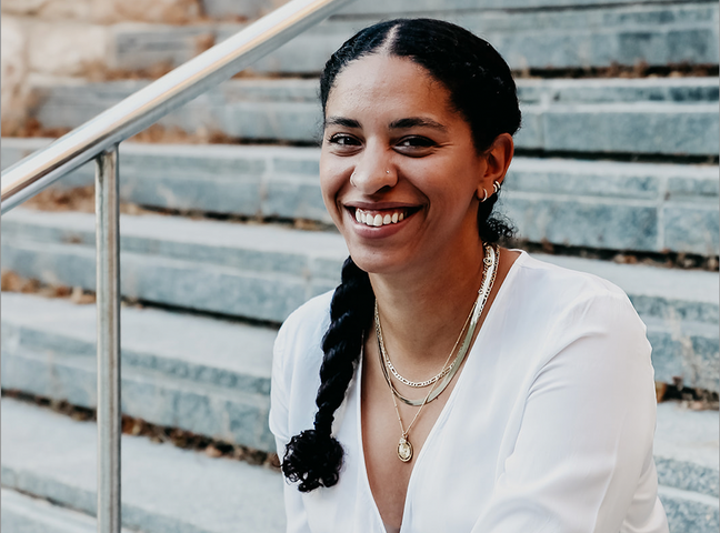 Natalya Mason wearing a white shirt and necklaces sitting on outdoor steps smiling into the camera