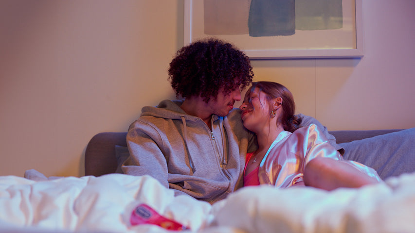 Au lit, deux personnes s’apprêtent à s’embrasser, une bouteille de lubrifiant Durex floutée à leurs côtés.
