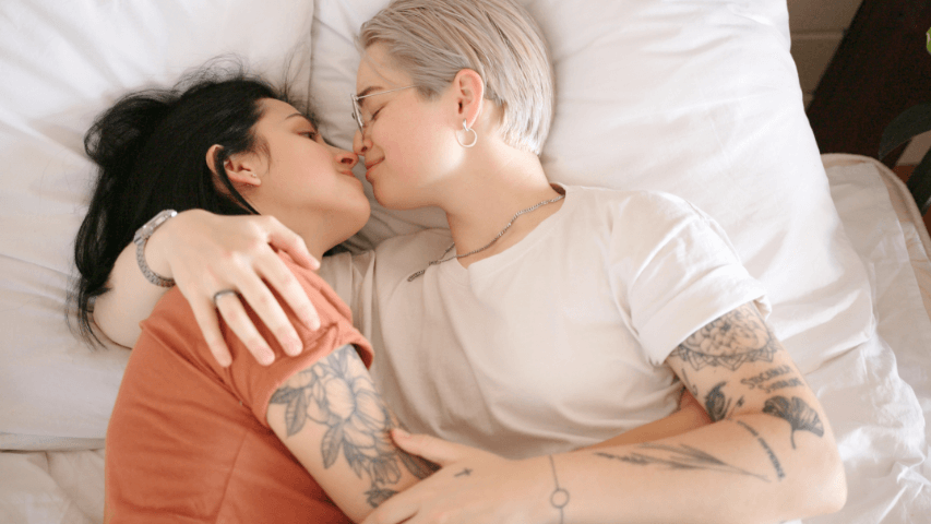 Two partners with tattoos rubbing noses in bed together.