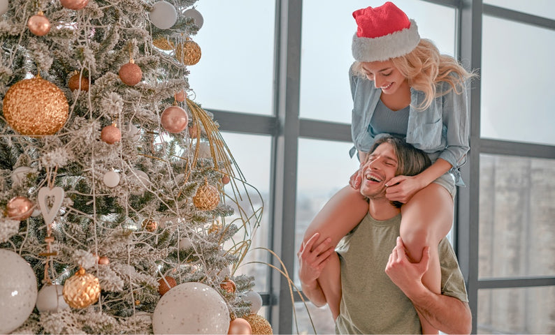 Une femme perchée sur les épaules d’un homme devant leur sapin de Noël.