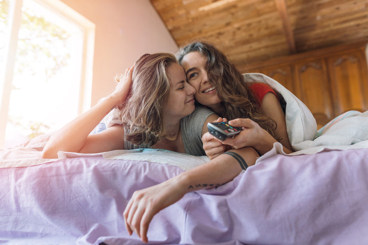Deux femmes blotties sous une couverture dans un lit et utilisant une télécommande.
