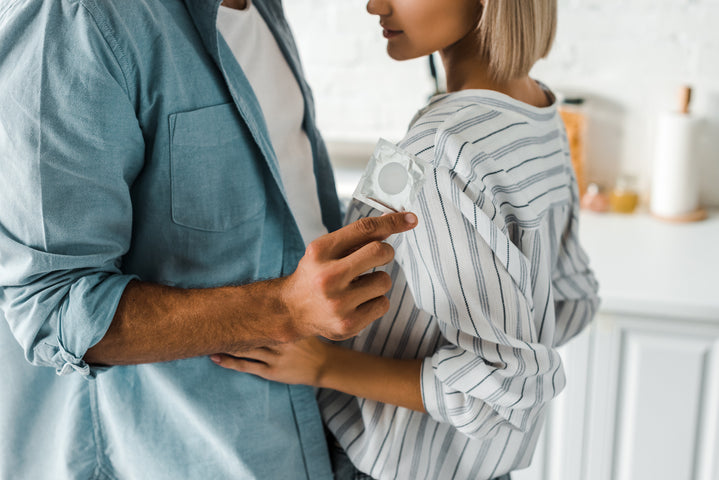 Un homme tenant un condom emballé devant l’appareil photo alors que sa partenaire s’approche de lui.