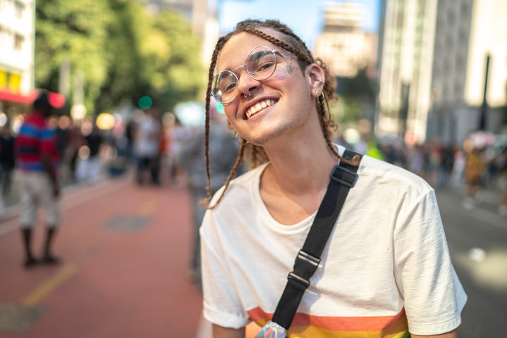 Une personne de la communauté LGBTQ+ avec des lunettes, des tresses et des brillants sur le visage souriant à l’appareil photo.