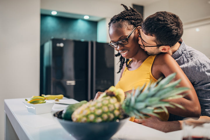 Man kissing his partner's neck while in the kitchen in front of a large pineapple