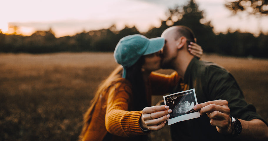 Couple kissing in a field while holding out a photo of their ultrasound baby for the camera