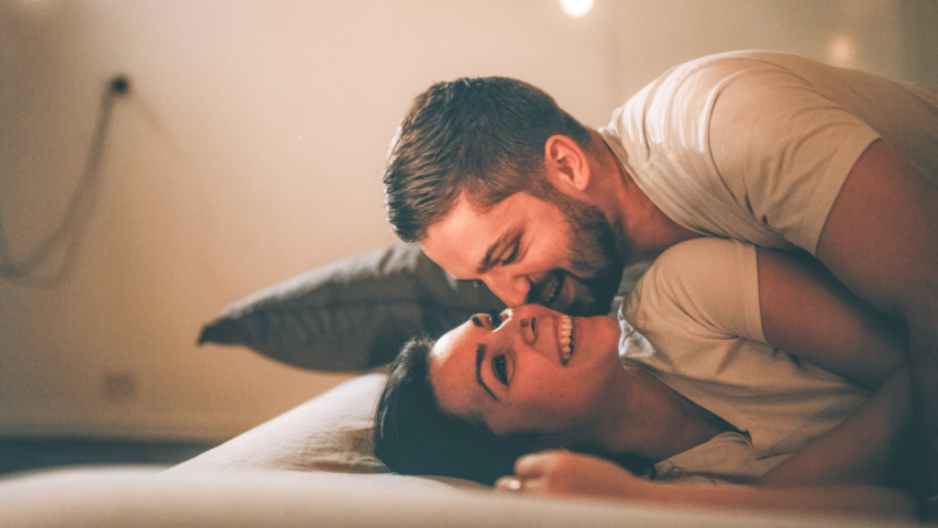 Man coming closer to his partner’s face while she smiles below him in bed.