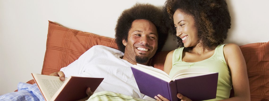 Couple looking at each other and smiling while reading separate books in bed.