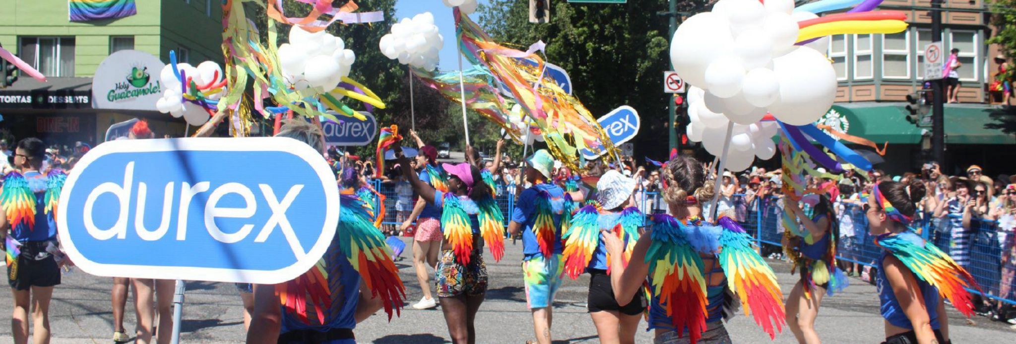 Image of a group of people wearing rainbow wings and carrying balloons and Durex signs walking in a Pride parade.