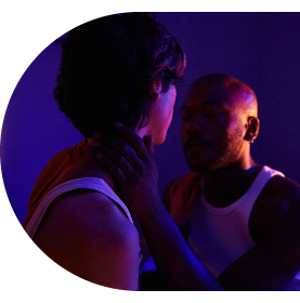 Man reaching for his partner’s neck about to kiss under fuchsia and sunset lighting.