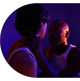 Un homme tend la main vers le cou de son partenaire pour l’embrasser sous un éclairage de crépuscule de couleur fuchsia