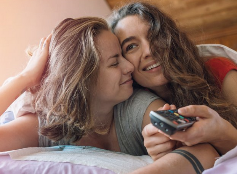 Deux femmes sous les couvertures qui se cajolent et sourient en tenant une télécommande.