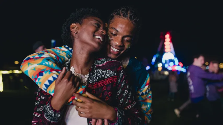 Two people wearing vibrant patterned clothing holding each other and laughing on a lit walkway at night.