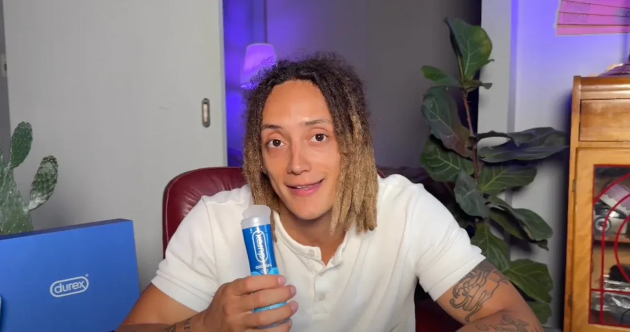 Le sexpert Durex Esteban Palacio tient une bouteille de lubrifiant Durex Chatouillement en parlant à la caméra; une boîte Durex bleue et des plantes sont visibles derrière son bureau.